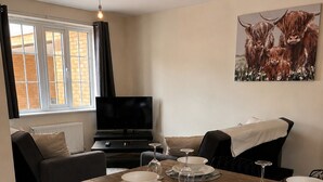 Un'area pranzo con un lungo tavolo di legno apparecchiato per quattro persone, una televisione sullo sfondo e un quadro raffigurante bestiame delle Highlands appeso al muro.