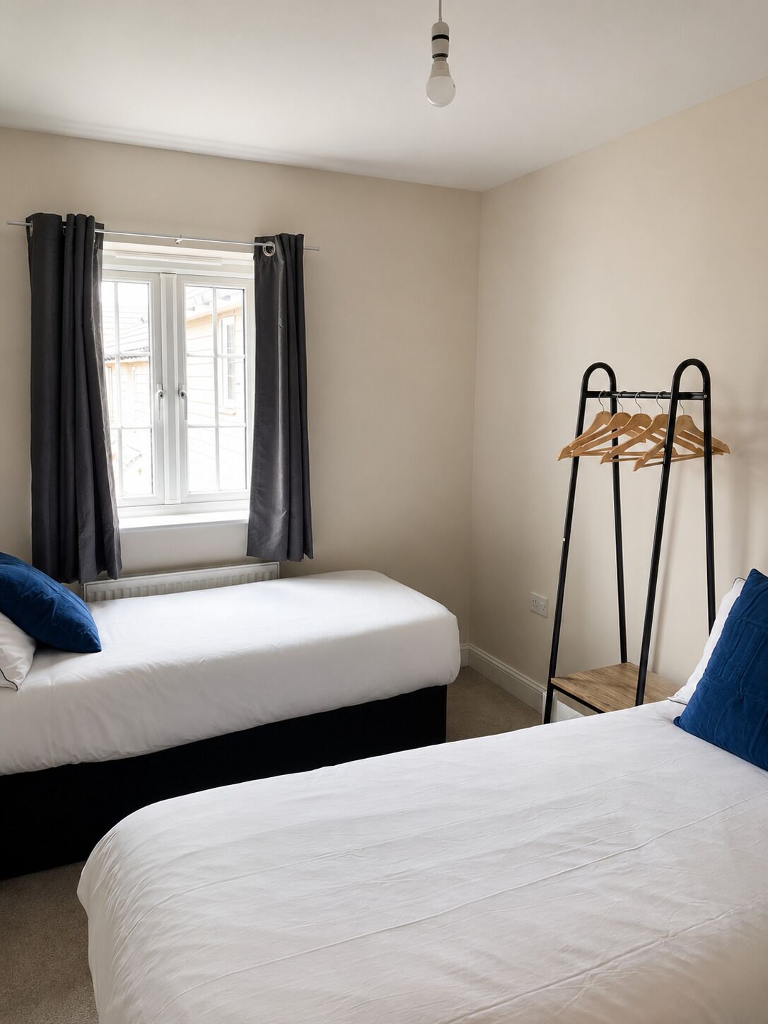 Una habitación con dos camas, una ventana con cortinas, un perchero y una bombilla.