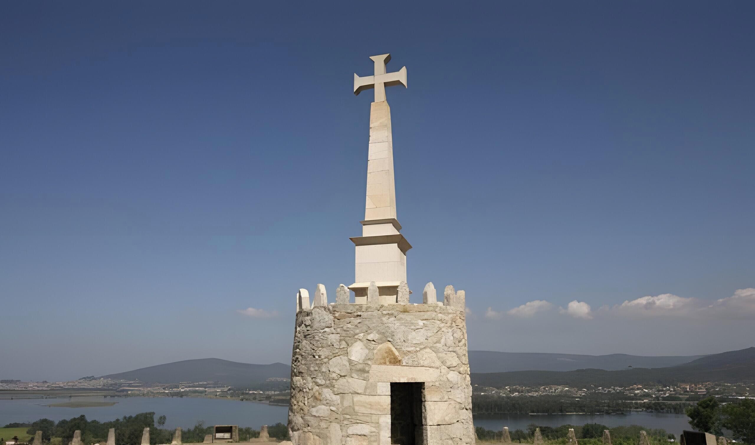 Una torre de pedra amb una creu al capdamunt, amb vista a una massa d'aigua i un paisatge urbà allunyat.