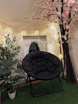 A black chair in a room with artificial greenery, a wall-mounted window, and string lights.