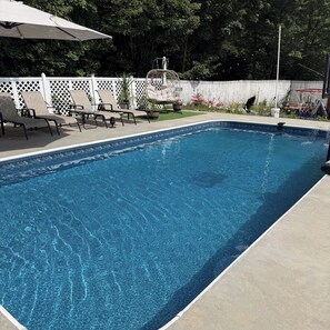 A rectangular swimming pool with a clear blue water surface, surrounded by a white fence, and equipped with lounge chairs and a table.