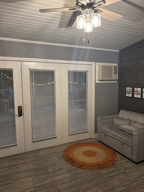 A room with a ceiling fan, a grey sofa, a patterned rug, and a wall-mounted air conditioning unit.