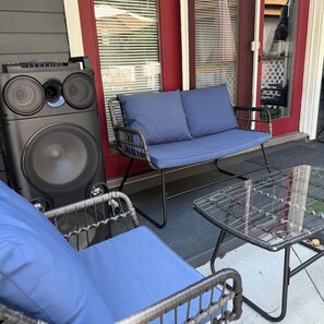 Eine Terrasse mit einem blauen Sitzmöbel, einem Korbstuhl und einem Glastisch.