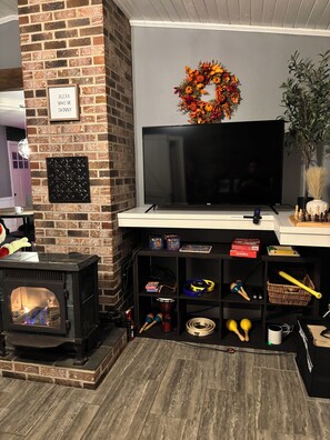 A living room with a brick fireplace, a flat-screen TV, and shelves with board games. - Lake George Getaway w/ Private Pool / Sleeps 12  3BR (Athol)