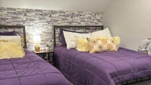 Quarto com duas camas, um bean bag cinza, uma mesinha de cabeceira com abajur e um tapete cinza.