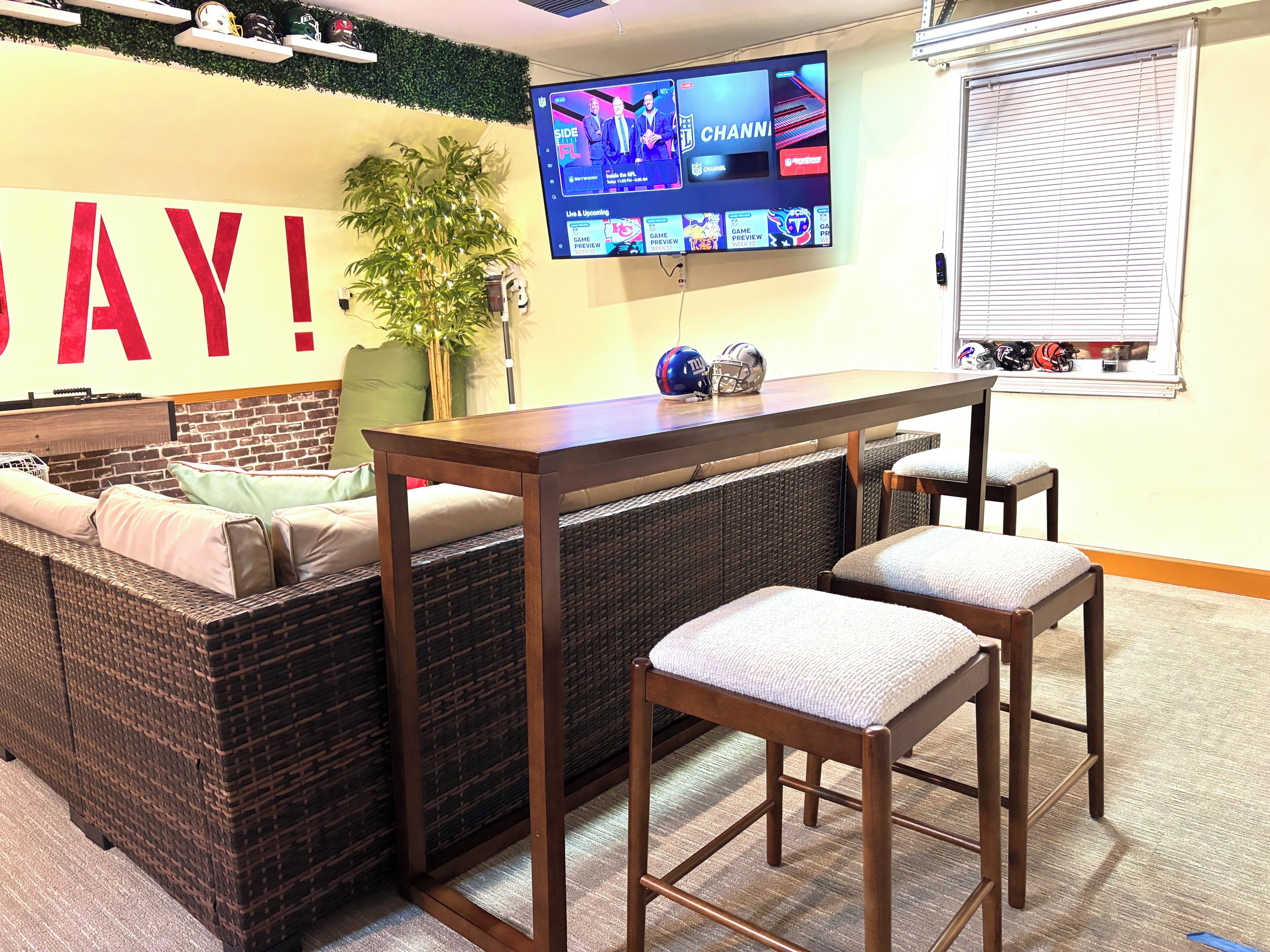 A room with a large TV displaying sports content, a sofa with cushions, a small table, and a window with blinds.