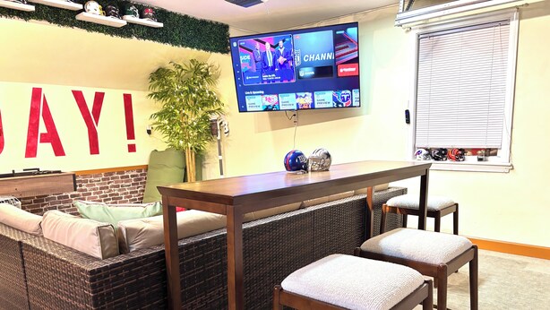 Quarto com uma televisão grande exibindo conteúdo esportivo, um sofá com almofadas, uma mesa pequena e uma janela com persianas.