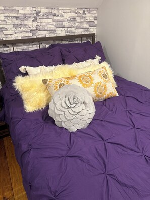 A bed with a purple bedspread, a fluffy white pillow, and decorative pillows with floral patterns.