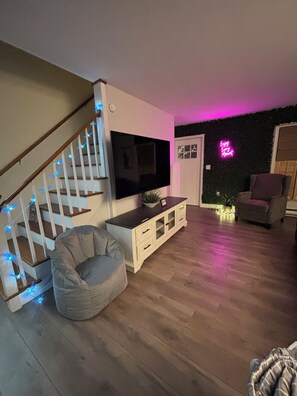 A modern living room with a staircase, a flat-screen TV, a bean bag chair, and a neon sign on the wall. - Lake George Getaway w/ Private Pool / Sleeps 12  3BR (Athol)