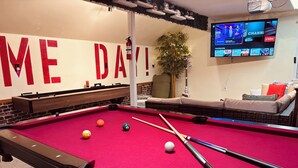 Uma mesa de bilhar com bolas e tacos, um sofá, uma televisão exibindo esportes e uma parede com texto grande.