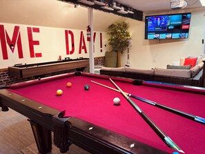 Ein Billardtisch mit Kugeln und Spielstöcken, eine Sitzecke mit Sofa, ein Fernseher, der Sport zeigt, und eine Wand mit großer Schrift.