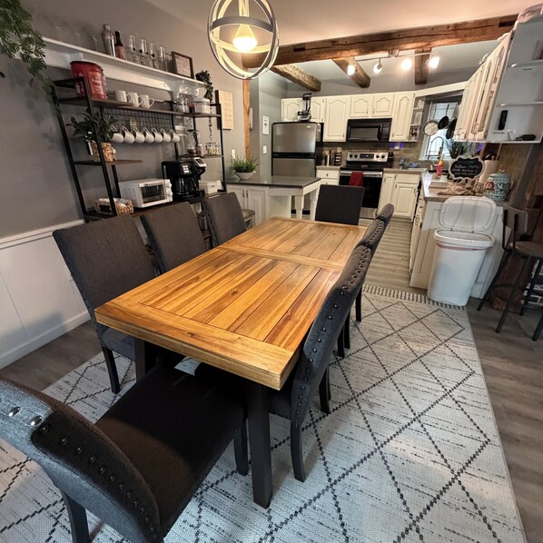 A dining area with a wooden table and chairs, kitchen in the background, and a patterned rug on the floor.