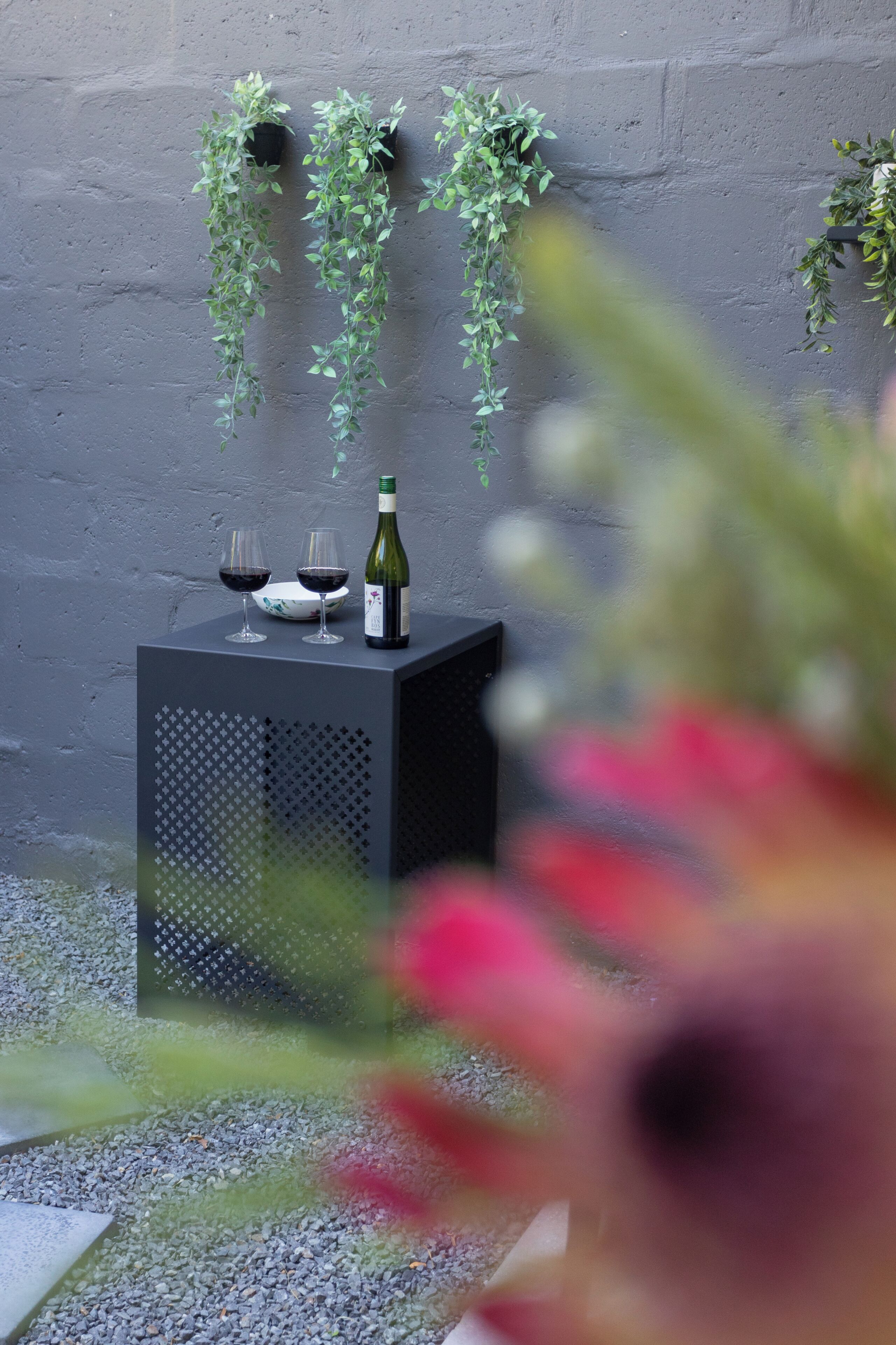 A small table with a bottle and glasses, surrounded by greenery against a textured wall.