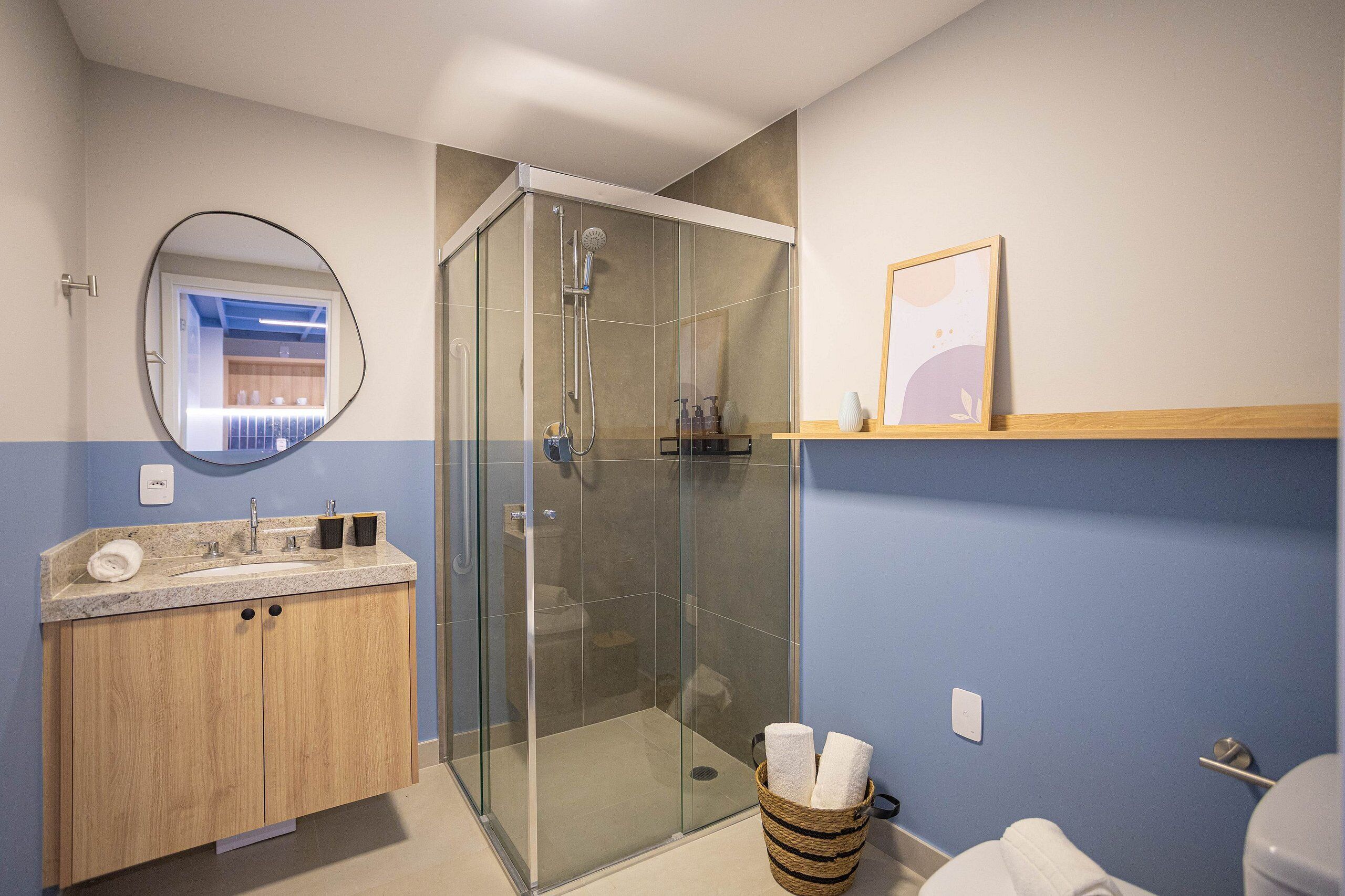 A bathroom with a glass-enclosed shower, a wooden vanity, a round mirror, and a framed picture on the wall.