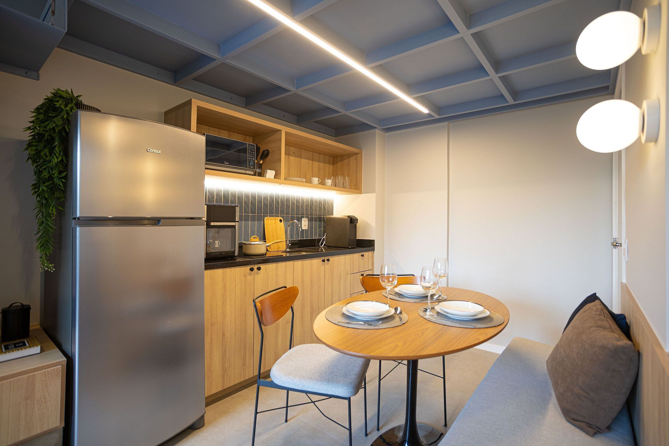 Cocina compacta con electrodomésticos modernos, comedor con mesa y sillas redondas, y techo con vigas a la vista.