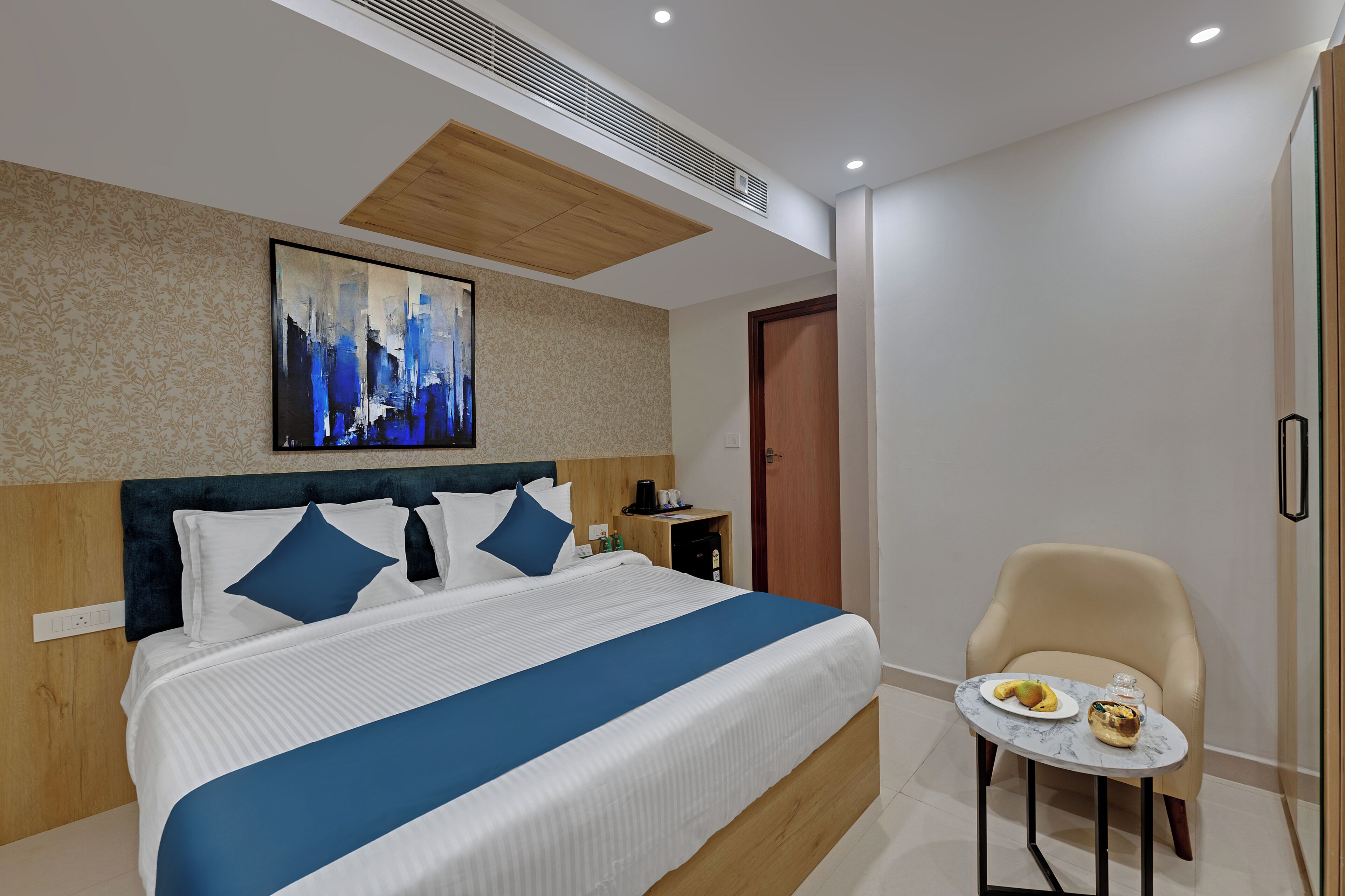 A modern hotel room with a large bed, a small table with fruit, a chair, and a painting on the wall.