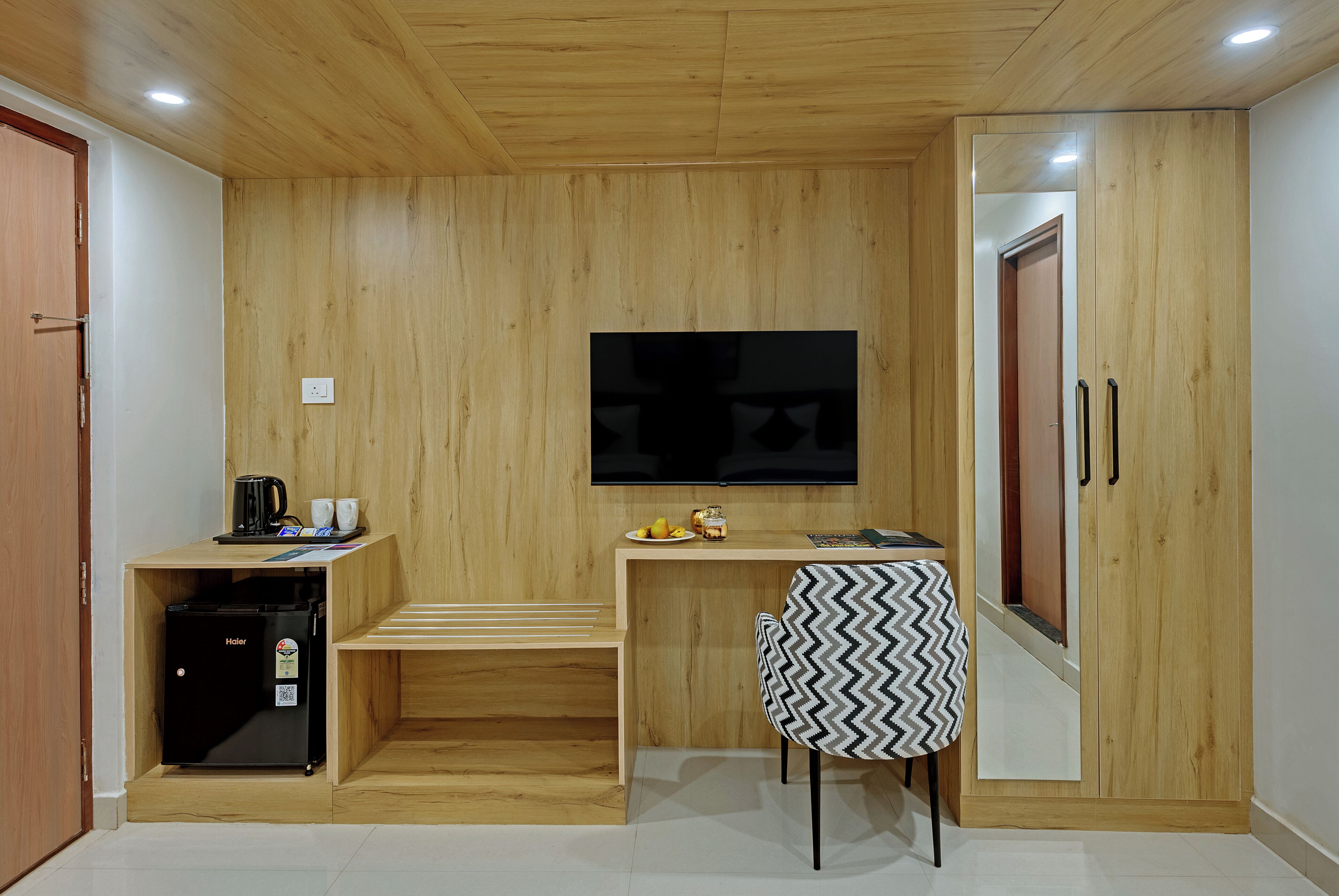 A compact room with a wooden wall, a flat-screen TV mounted above a small table, a refrigerator, and a chair with a patterned cover.