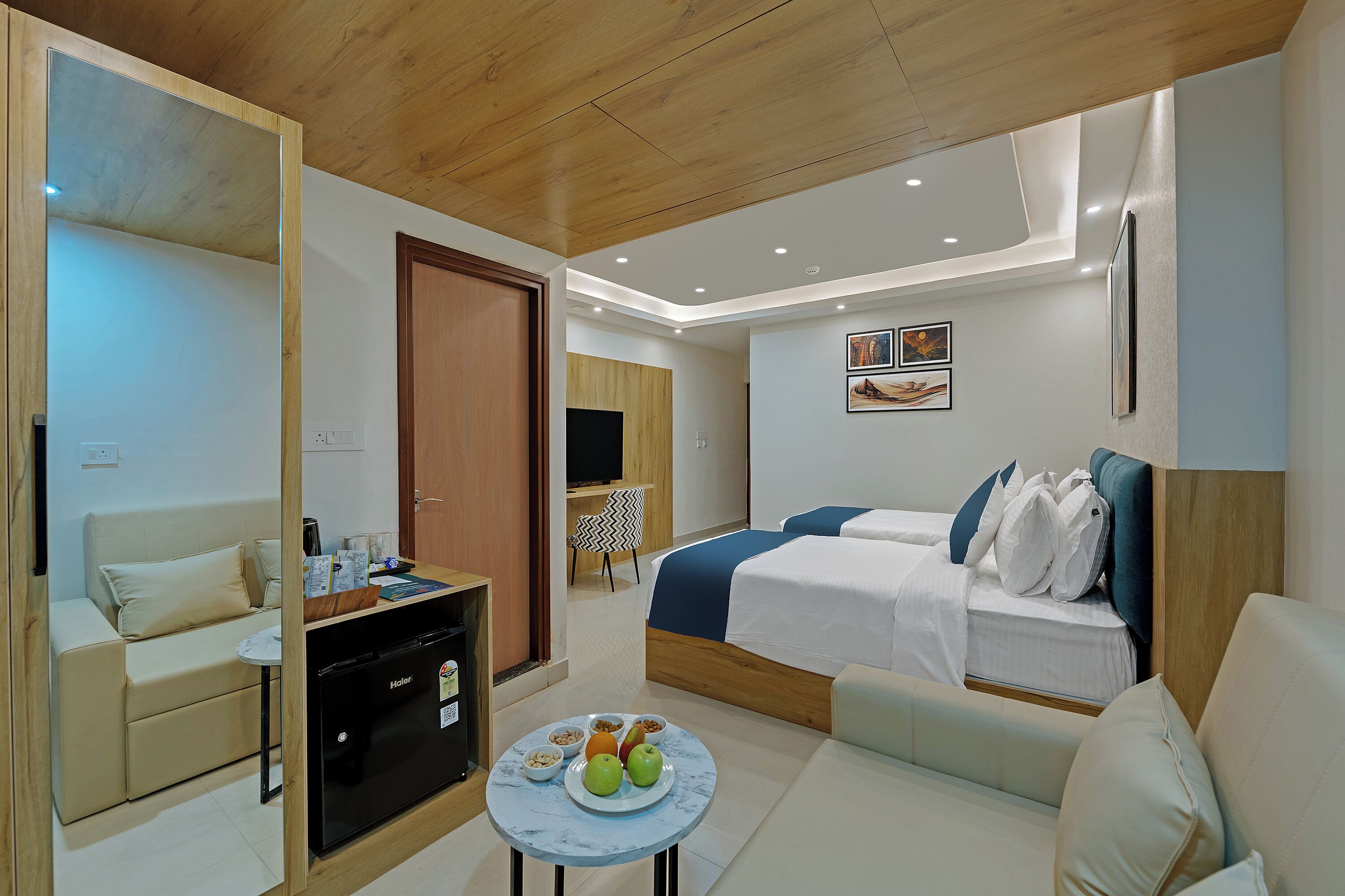 A hotel room with a bed, a sofa, a desk with a coffee machine, a small table with fruit, and a mirror.