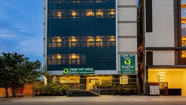 Un moderno edificio de hotel con fachada de vidrio, señalización que dice 'Quality Inn Bhavishya' y una entrada visible con escalones.