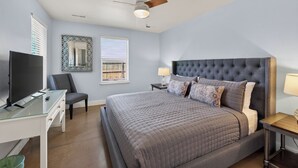 Une chambre à coucher comprenant un lit, un bureau, une chaise, un téléviseur, un ventilateur de plafond et une fenêtre avec des stores.
