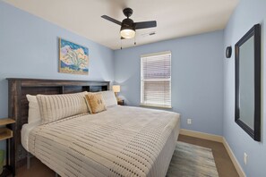 Une chambre à coucher avec un lit, un ventilateur de plafond, une fenêtre avec des stores, un tableau au mur et un miroir encadré.