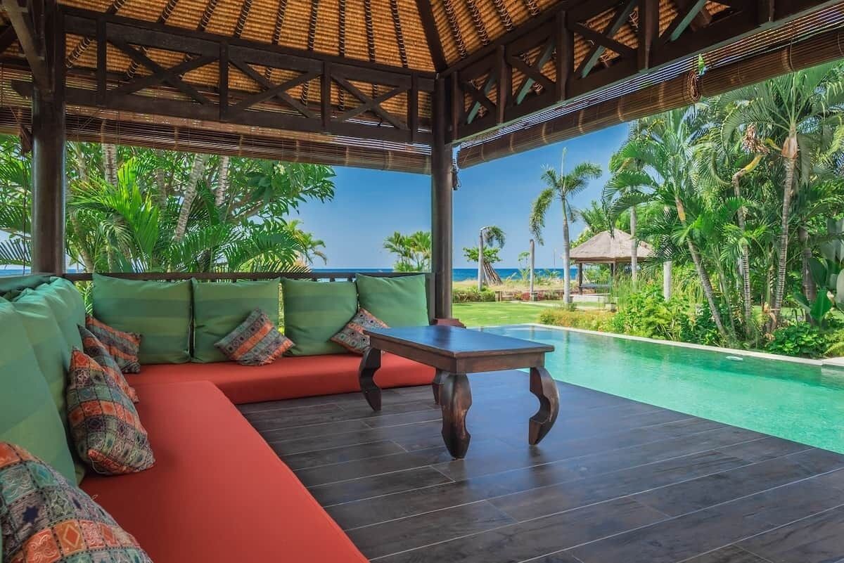 Un área de estar al aire libre y techada, con piscina, rodeada de abundante vegetación y vista despejada al océano.