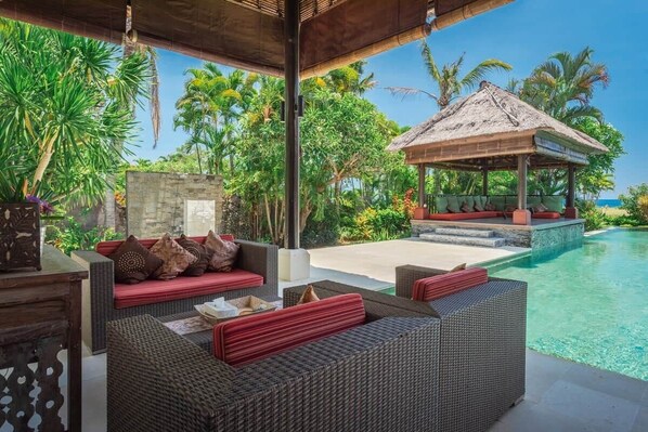A covered outdoor seating area with wicker furniture and red cushions, overlooking a pool and tropical vegetation.