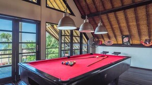 Uma mesa de bilhar com bolas, iluminação pendente e vista através de grandes janelas.