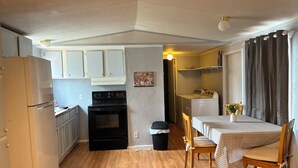 Eine kompakte Küche mit Kühlschrank, Herd und Waschmaschine. Ein Esstisch mit Stühlen und ein kleines Fenster mit Vorhängen sind ebenfalls vorhanden.