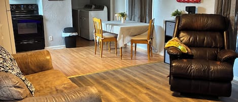 Una sala de estar con sofá de cuero, sillón reclinable marrón, mesa de comedor con sillas, ventilador de techo y una cocina al fondo.