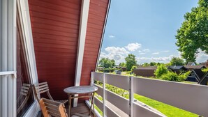 Un balcón con una silla de madera, una mesita y vistas a un barrio residencial con casas y vegetación.