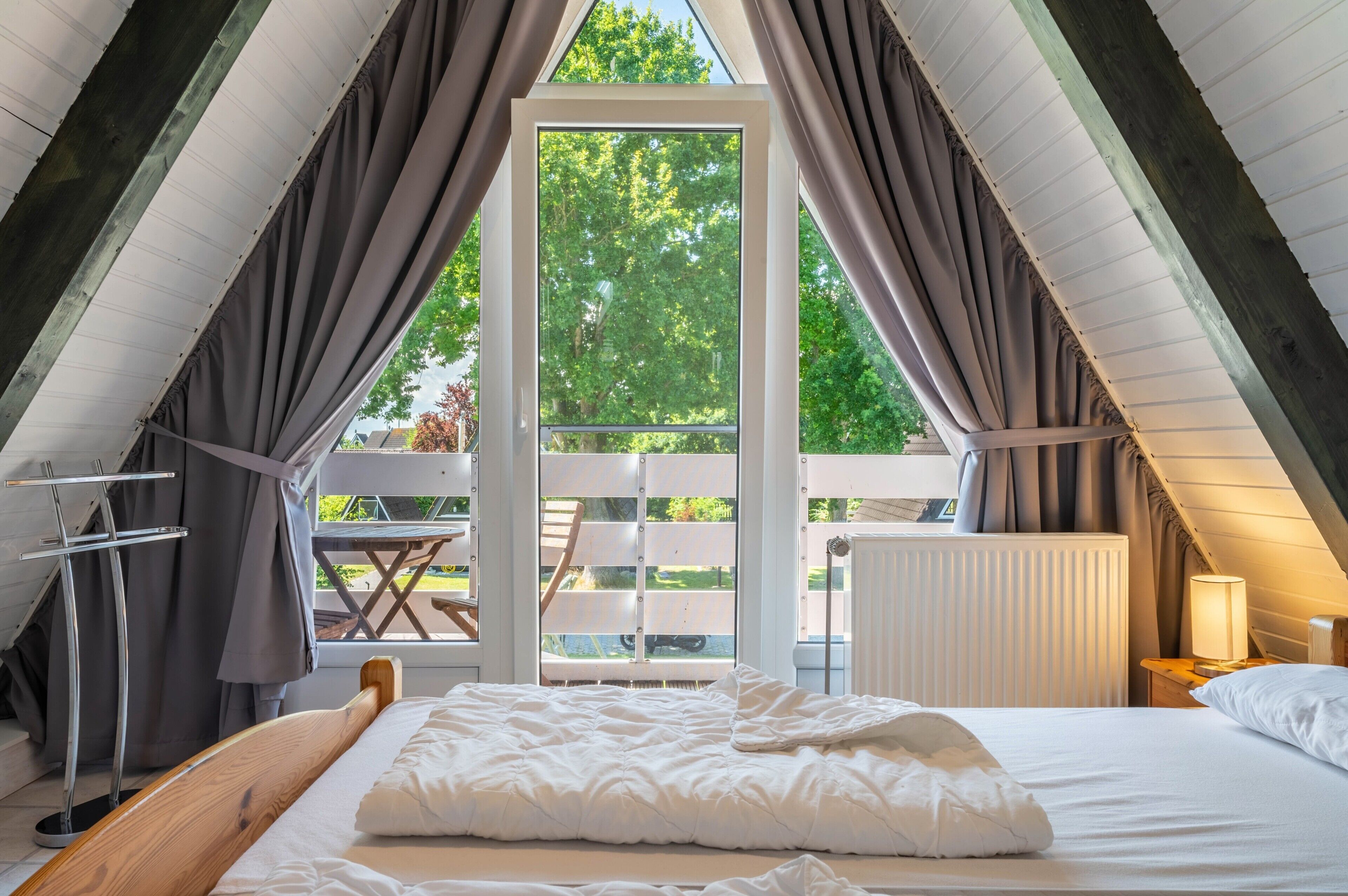 An attic bedroom with a large window, a wooden bed, a table, and a chair.