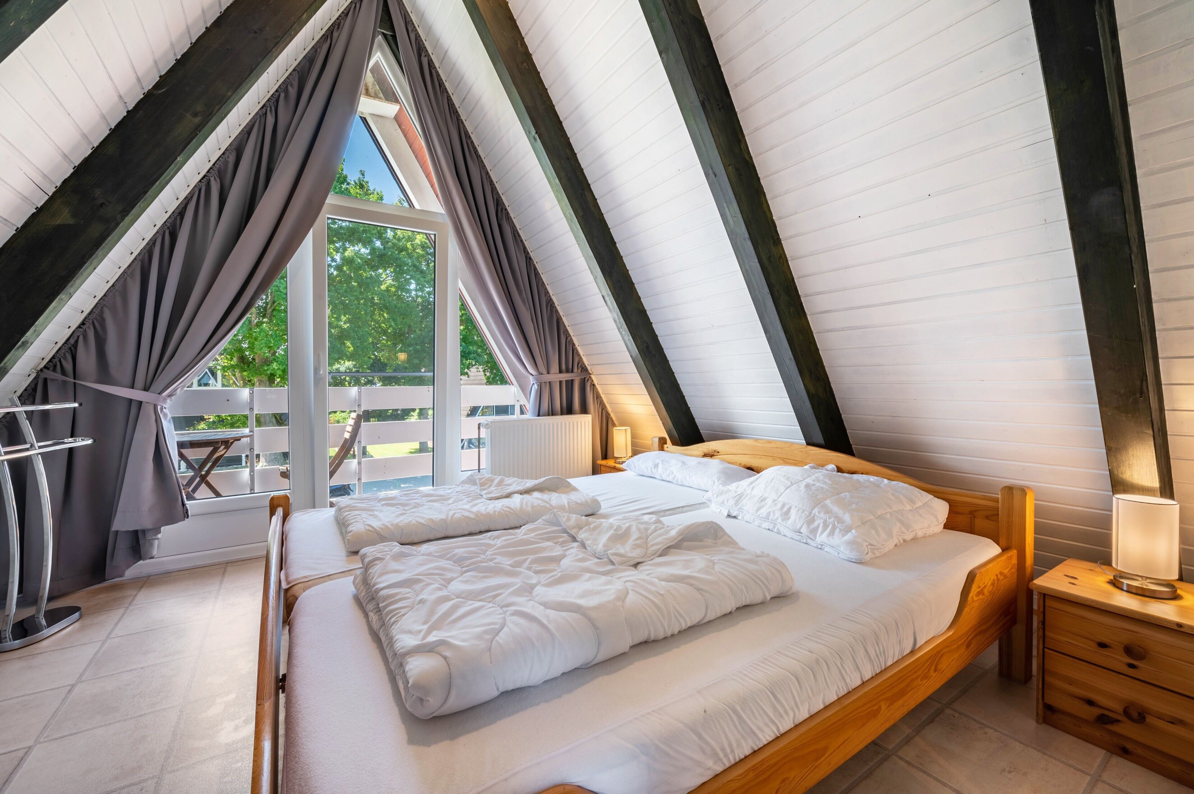 A bedroom with a sloped ceiling, a bed with white bedding, a nightstand, and a window with curtains.