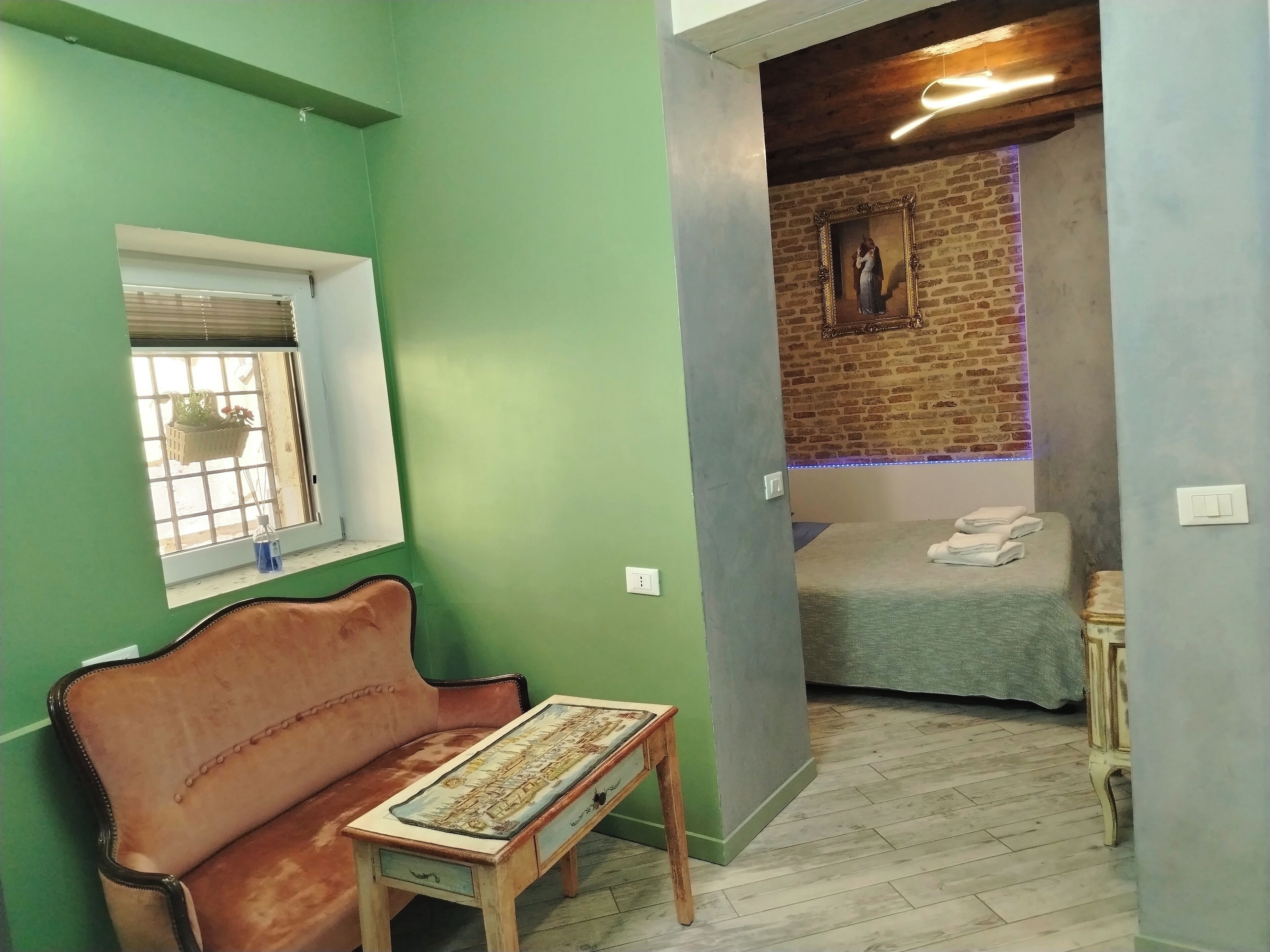 A room with a green wall, a window with blinds, a brown leather sofa, a small wooden table, a bed with a blue blanket, and a brick wall with a framed picture.