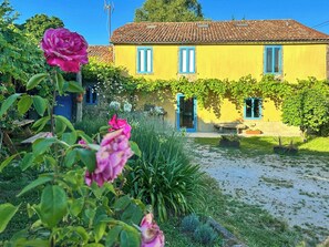 A yellow building with a tiled roof, surrounded by a garden with pink flowers and green plants. - Galician stone house with hórreo and garden (Bustelo)