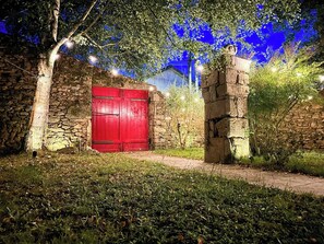 Ein Steinhaus mit roter Tür, umgeben von Grün und beleuchtet mit Lichterketten.