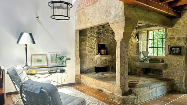 A room with a stone fireplace, a chair, a lamp, and a view of greenery outside. - Galician stone house with hórreo and garden (Bustelo)