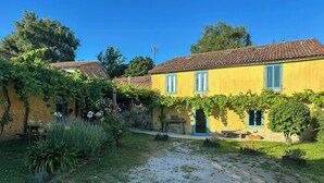 A yellow building with a tiled roof, surrounded by greenery and a gravel path. - Galician stone house with hórreo and garden (Bustelo)
