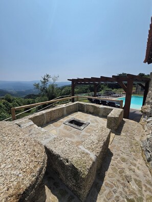 A stone fire pit with a pool and lounge chairs in the background. - Vale do Luar House (Celorico de Basto)