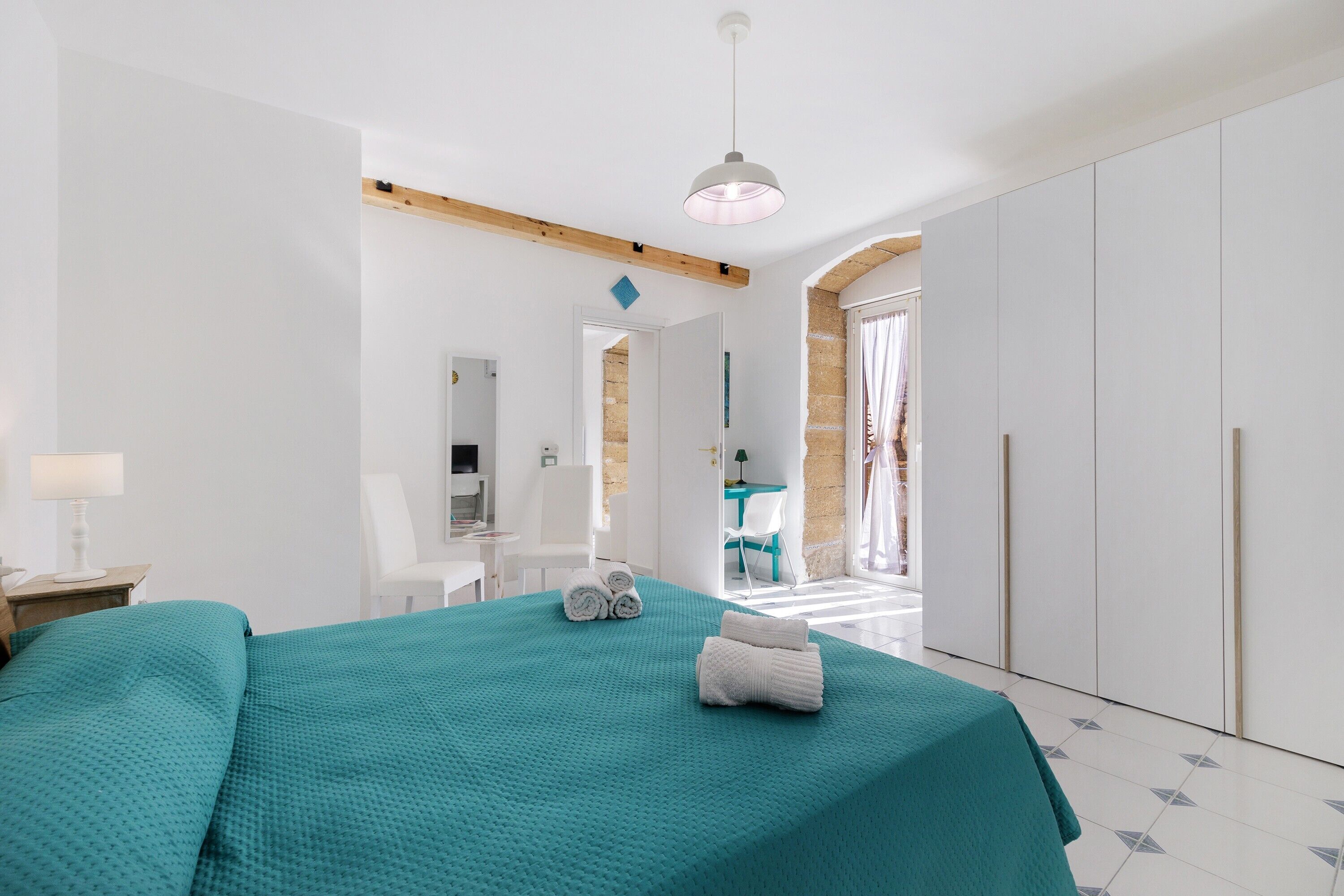 Quarto moderno com cama, escrivaninha e cadeira. É possível ver um guarda-roupa e um banheiro por uma porta aberta.
