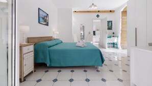 Un dormitorio con cama, mesitas de noche, un cuadro, una lámpara colgante y un baño visible a través de una puerta abierta.