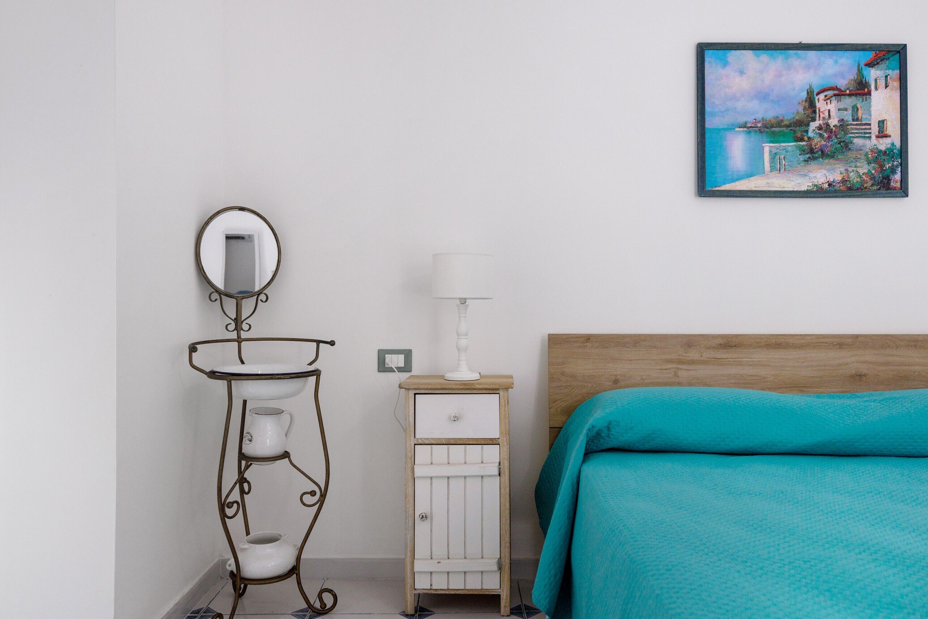 Una camera da letto con un letto di legno, un comodino bianco con una lampada, un piccolo armadietto bianco e uno specchio fissato alla parete.