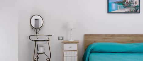 Un dormitorio con cama de madera, una mesita de noche blanca con lámpara, un armario pequeño blanco y un espejo montado en la pared.