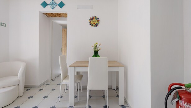 Una habitación con un sofá blanco, una mesa de comedor de madera con dos sillas y un pequeño florero verde con flores.