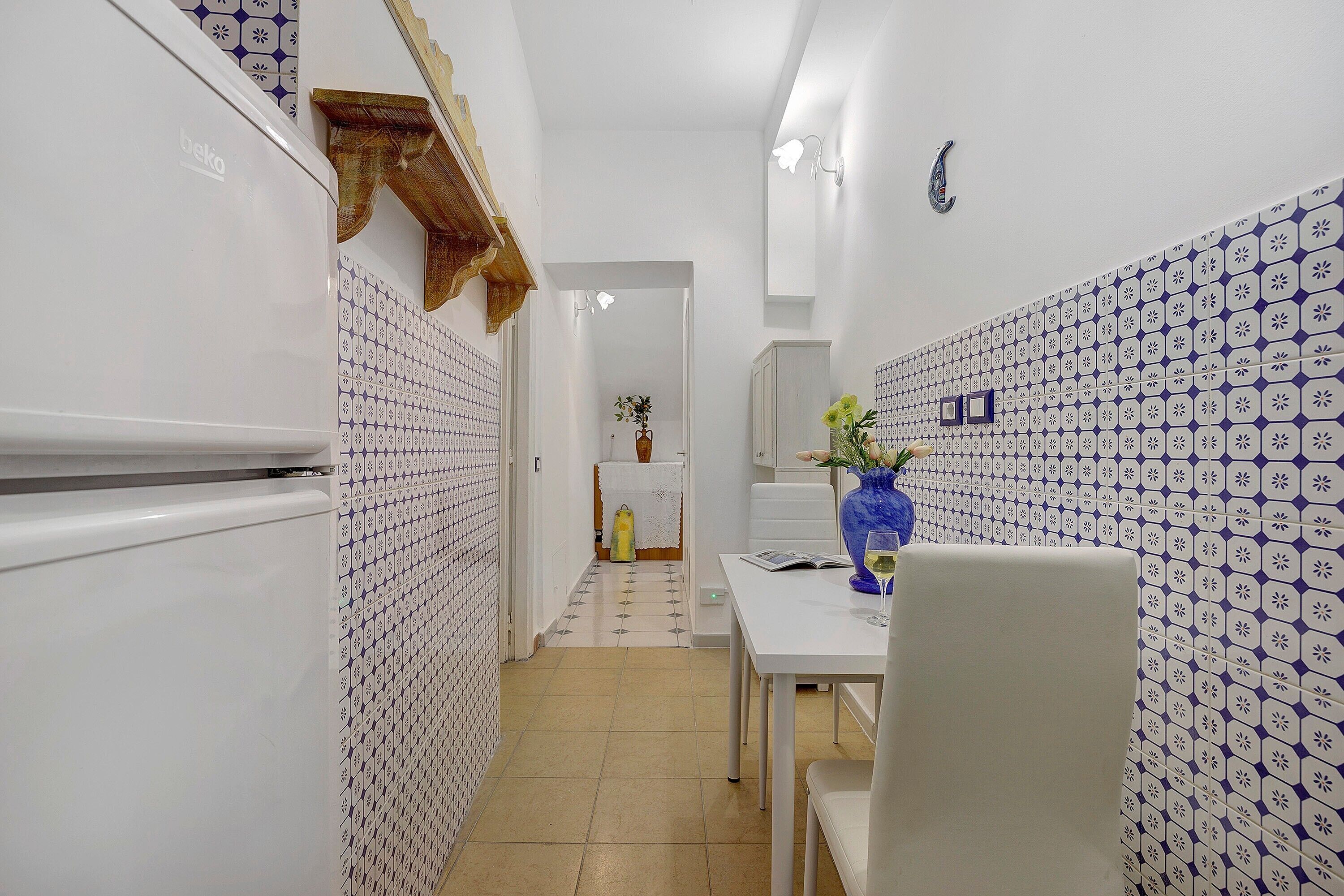 A narrow hallway with patterned walls, a dining area with a table and chairs, and a refrigerator on the left.