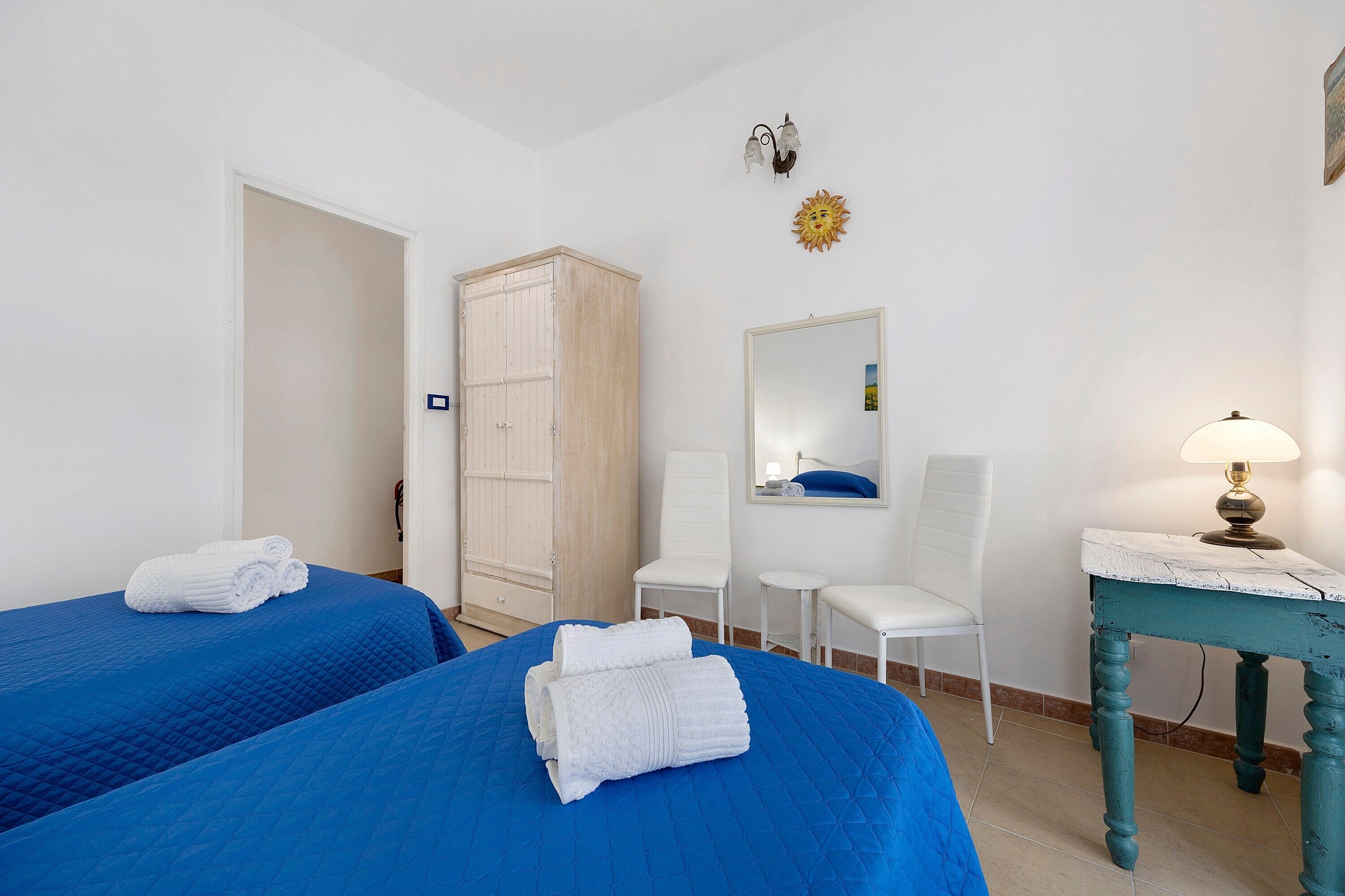 A hotel room with two beds, a wardrobe, a mirror, a chair, and a table with a lamp.