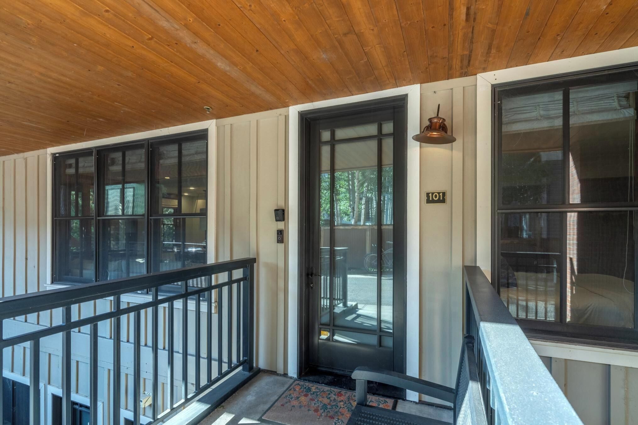 A covered porch with a wooden ceiling, a black railing, and a door with a number plate.