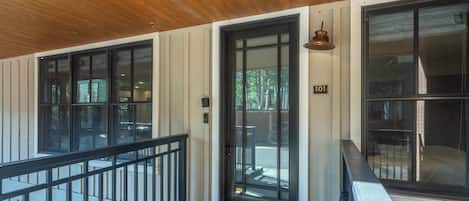 A covered porch with a wooden ceiling, a black railing, and a door with a number plate.