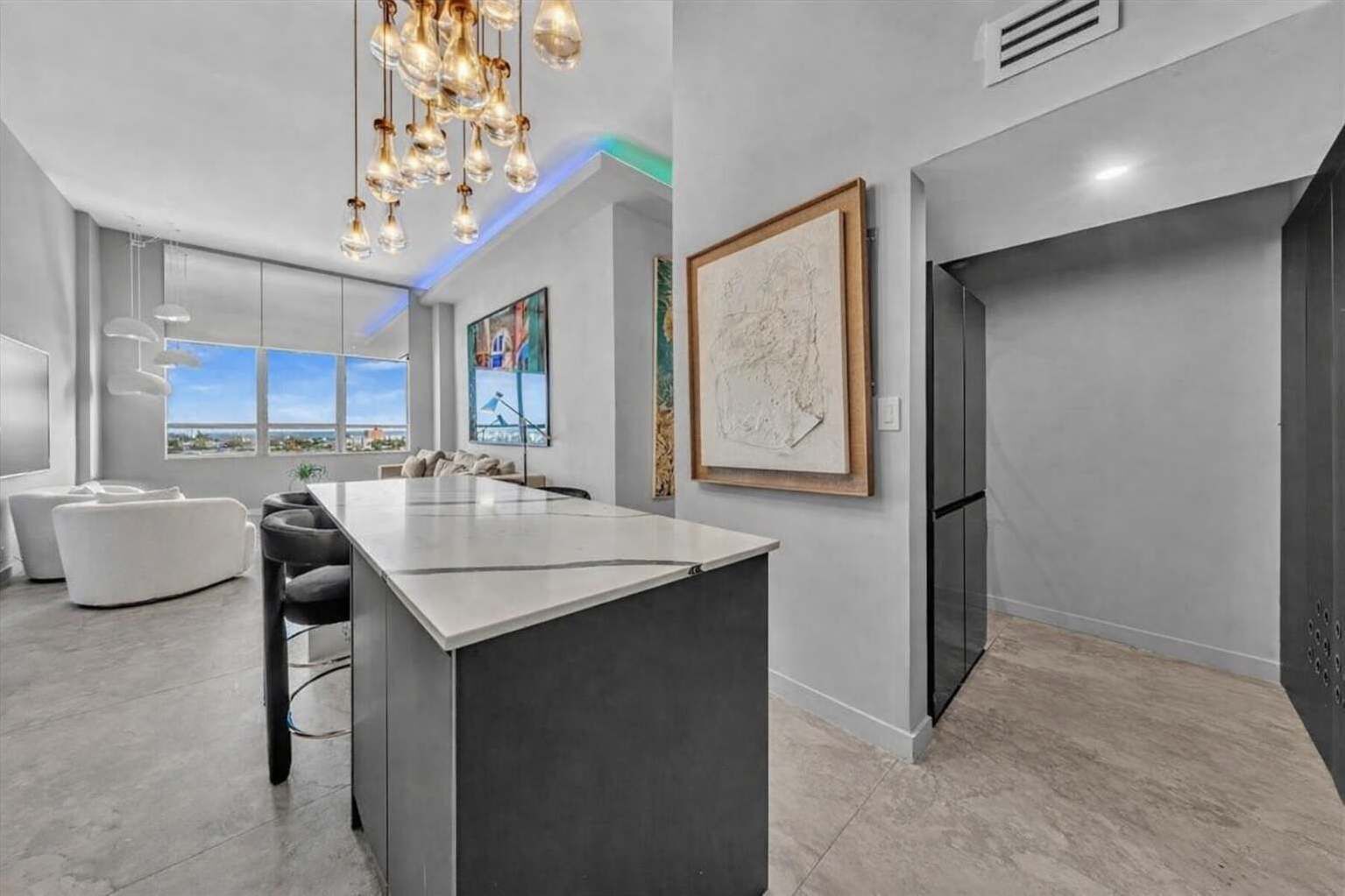 Chic open kitchen with a marble-topped island, bar seating, and a dazzling cluster of pendant lights above. LED accent lighting and skyline views through expansive windows tie the space to the stylish living area beyond.