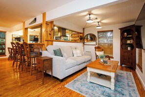 A spacious living area with a light-colored sofa, wooden dining table, and chairs. A kitchen is visible in the background with wooden cabinets and a hanging light fixture.